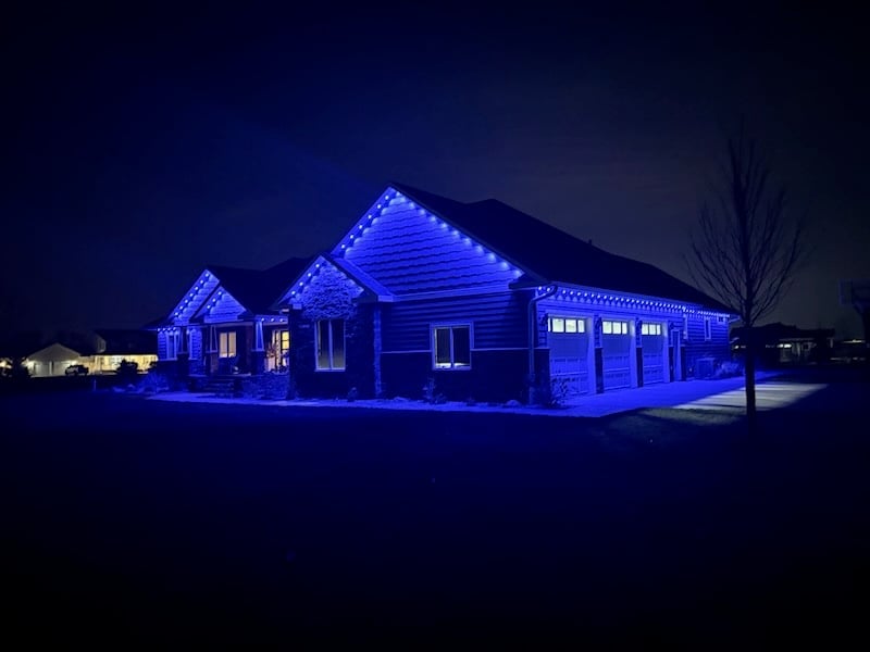 Modern house illuminated with bright blue LED lights at night, featuring a large garage and surrounded by dark landscape