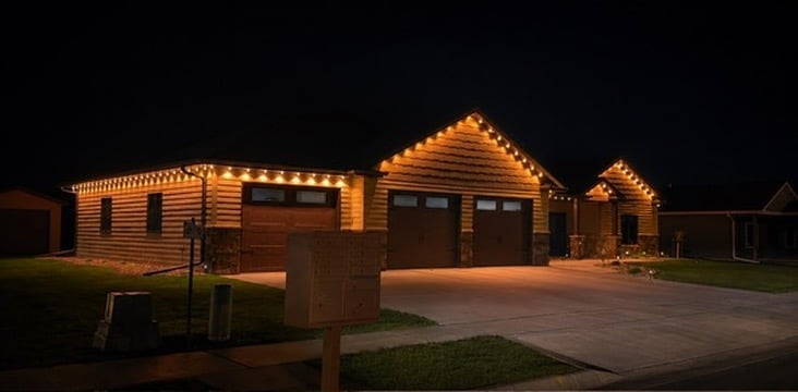 Nighttime view of illuminated wooden log cabin structures with warm lighting outlining the rooflines and walls against a dark sky.