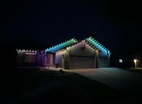 A residential house at night illuminated with colorful multicolored LED lights along the roofline and eaves against a dark sky
